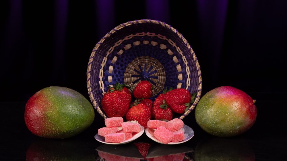 The foreground has a shimmering gummy with sugar on it and in the background there are strawberries and mangoes!