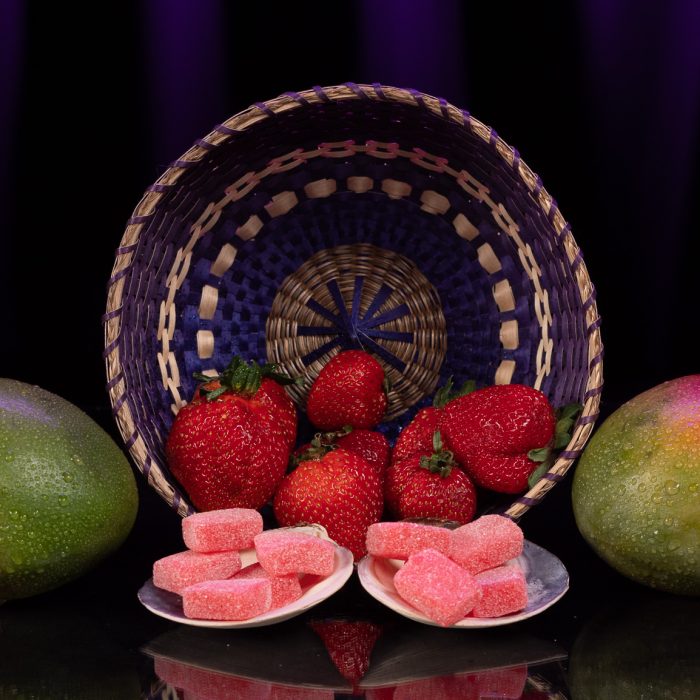 The foreground has a shimmering gummy with sugar on it and in the background there are strawberries and mangoes!